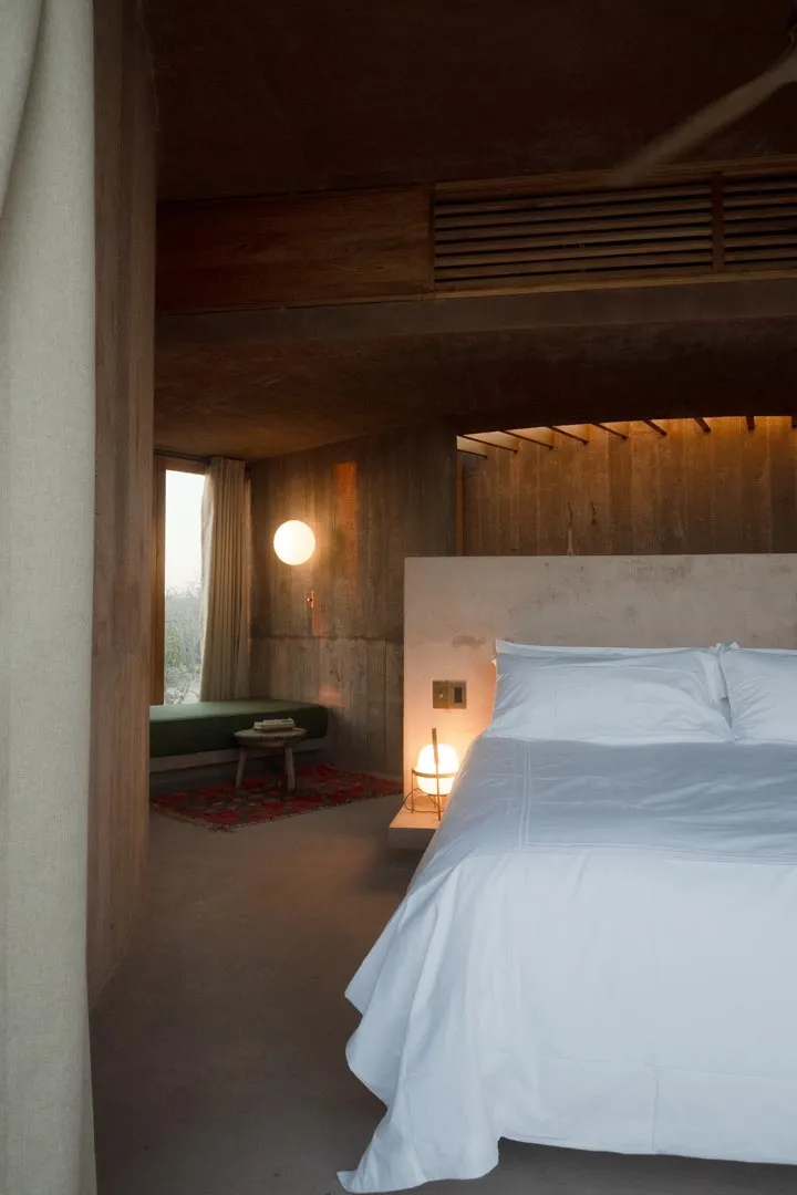 Suite interior at dusk: concrete walls, a low green daybed, and lamplight beside the bed